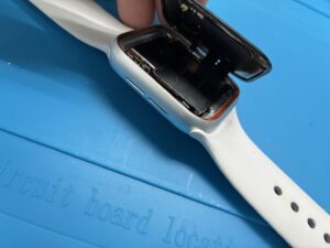 修理スタッフが指でApple Watch Series 3の画面を貝のように慎重に持ち上げ、内部を露出させている様子。黒い四角いバッテリーや、画面と基板を繋ぐ細い液晶ケーブル、フチのデリケートなセンサーが見える。